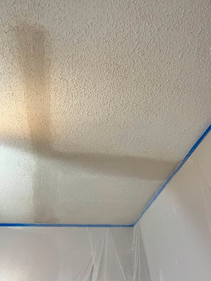 Popcorn ceiling mid-removal with blue tape and plastic masking protecting the walls